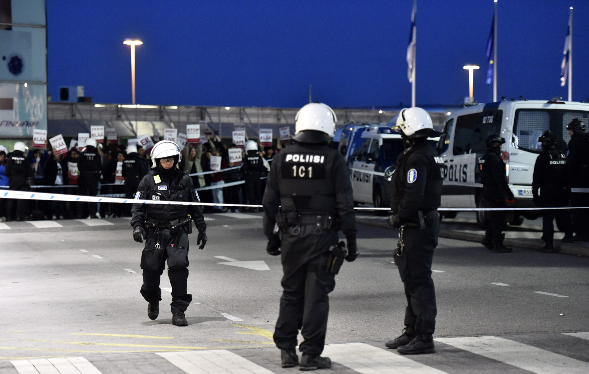 Tre poliser i skyddsutrustning med en demonstrerande folkmassa i bakgrunden.