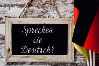A wooden-framed chalkboard with the question Sprechen sie Deutsch? do you speak German? written in German, and some flags of Germany against a rustic wooden background.