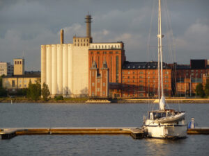 Vy över strandlinjen vid Academill, med brygga och segelbåt i förgrunden och Havtornen och silorna bortom vattnet.