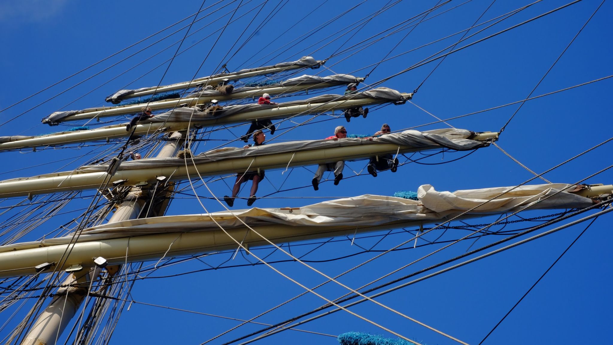 Flera personer klättrar i en enorm mast på segelskuta. Blå himmel som bakgrund.