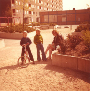 Förortsmiljö på 1970-talet. Två barn och en mamma på en grusgård framför ett höghus.