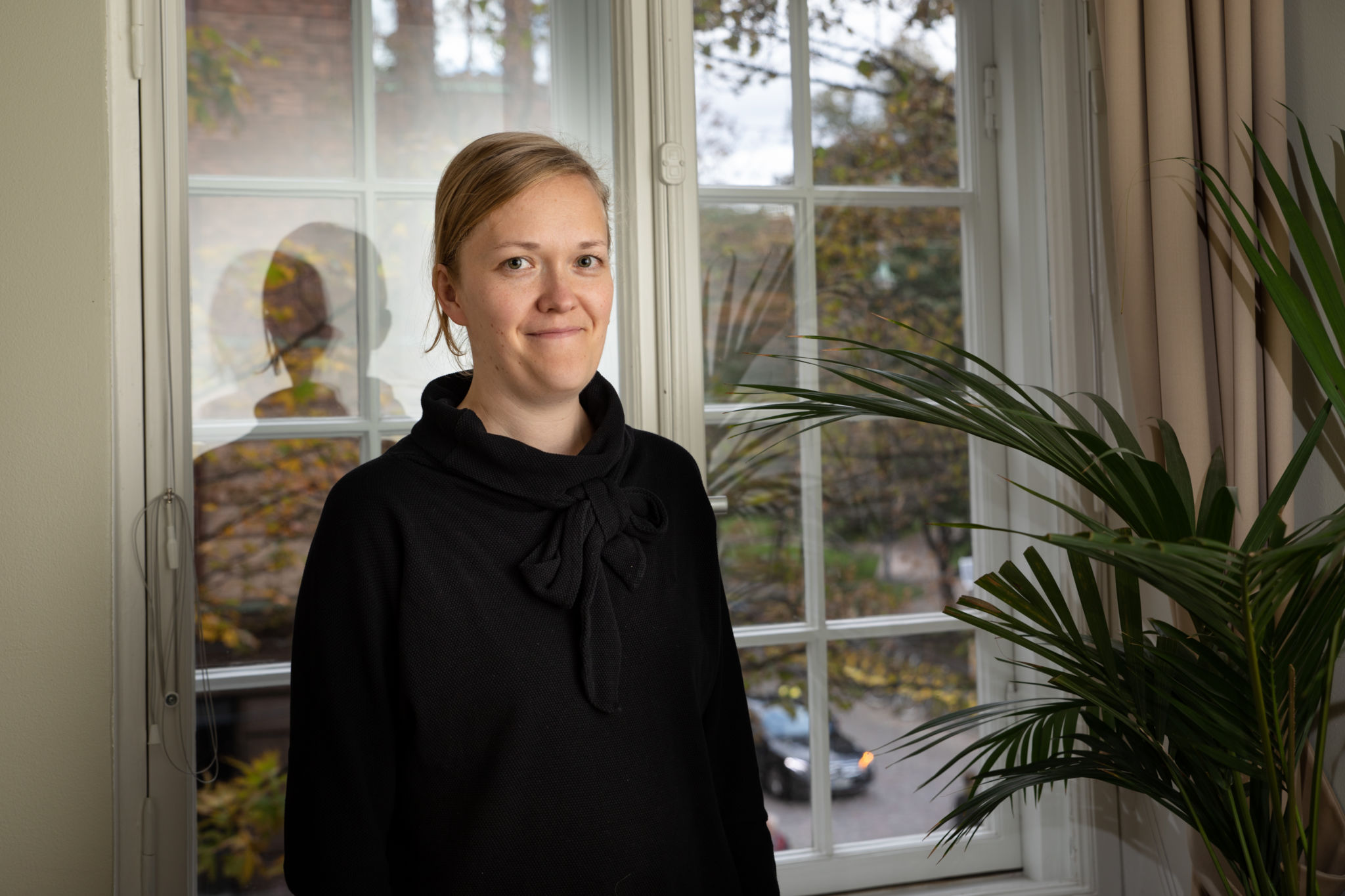Henni Alava klädd i svart, står framför ett fönster och en krukväxt.