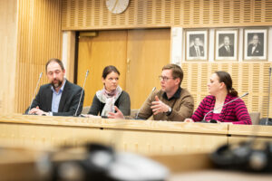 Fyra personer sitter bakom ett träfärgat podium och diskuterar under medborgarrådet i Korsholm. Svartvita porträtt i bakgrunden.