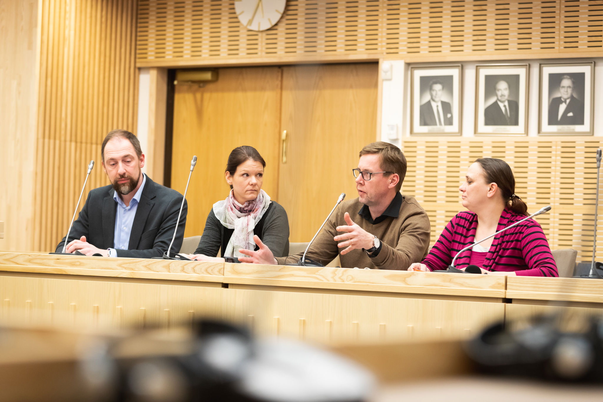 Fyra personer sitter bakom ett träfärgat podium och diskuterar under medborgarrådet i Korsholm. Svartvita porträtt i bakgrunden.