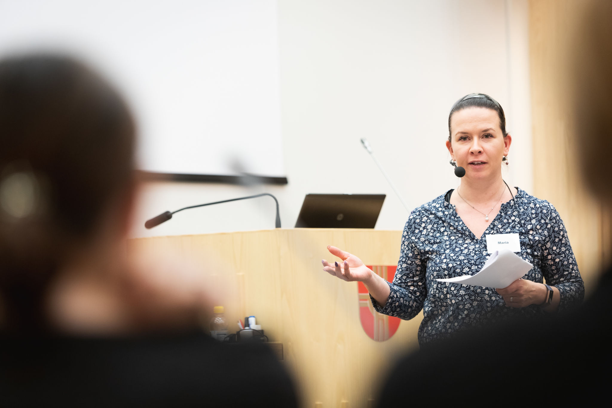 Maria Bäck med papper i hand och headset-mikrofon framför träfärgat podium och silhuetter av publikryggar i förgrunden.