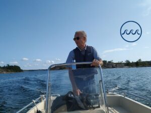 Professor Erik Bonsdorff kör en båt stående. I bakgrunden blått hav och blå himmel och uppe i höger hörn logon för det strategiska profilområdet Havet. Logon består av en cirkel med tre vågor i mitten.