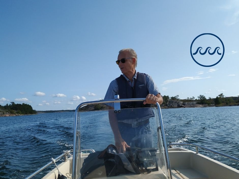 Professor Erik Bonsdorff kör en båt stående. I bakgrunden blått hav och blå himmel och uppe i höger hörn logon för det strategiska profilområdet Havet. Logon består av en cirkel med tre vågor i mitten.