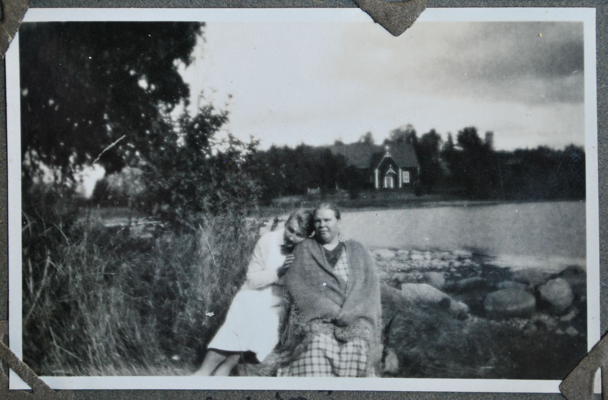 Svart-vit bild på två kvinnor där den ena lutar huvudet på den andras axel. I bakgrunden Själö kyrka.