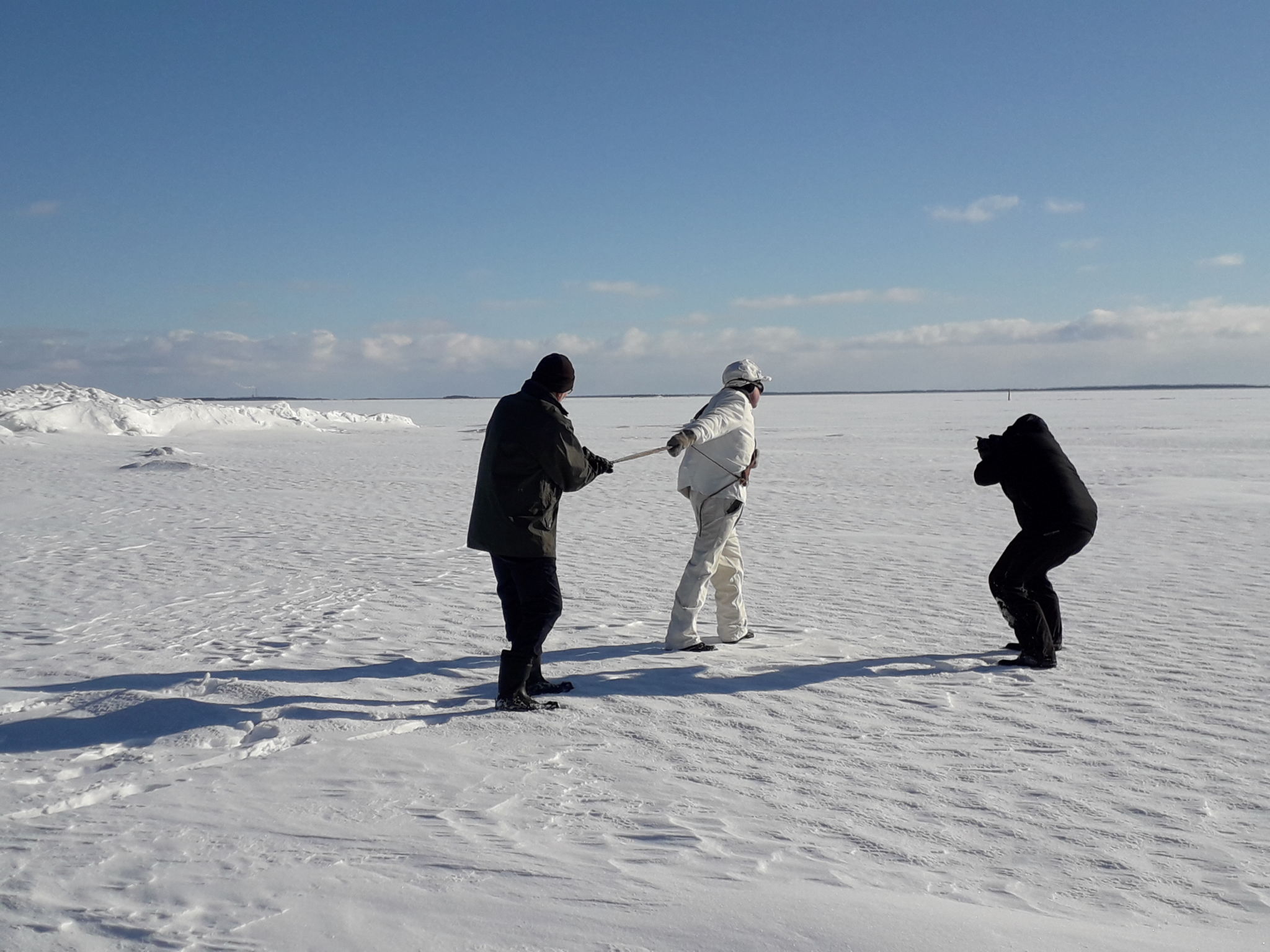 Två män går på isen på säljakt, en redje fotograferar dem.