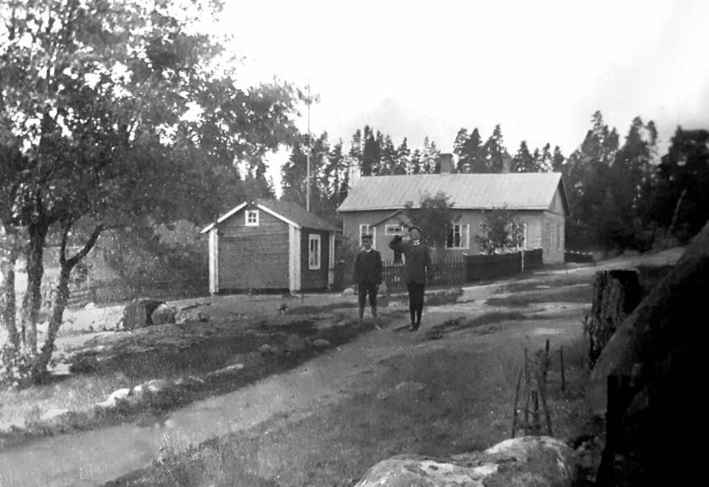 Två ganska otydliga personer på en väg framför ett större och ett mindre hus.