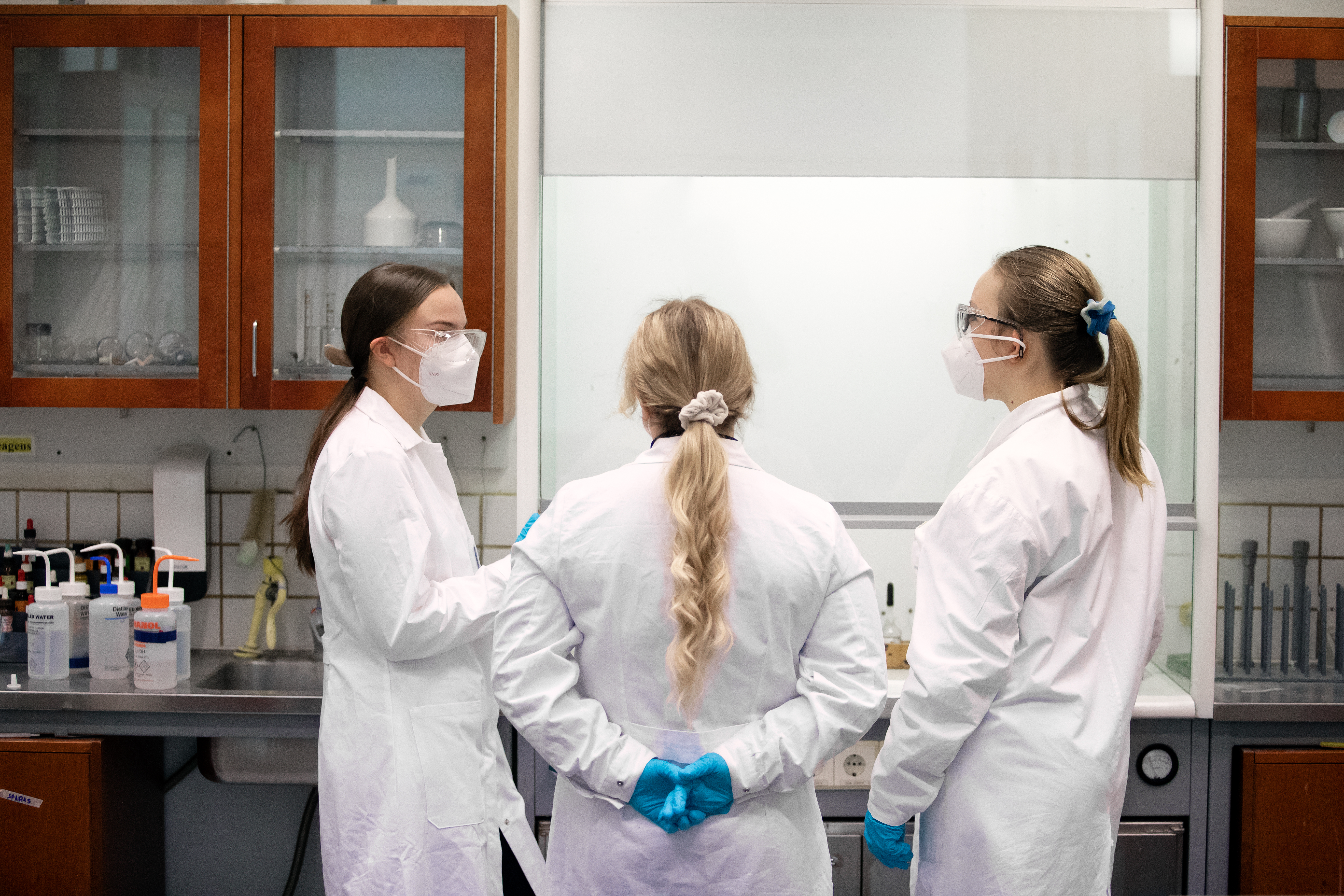 Tre studerande i labbrockar talar i ett laboratorium.