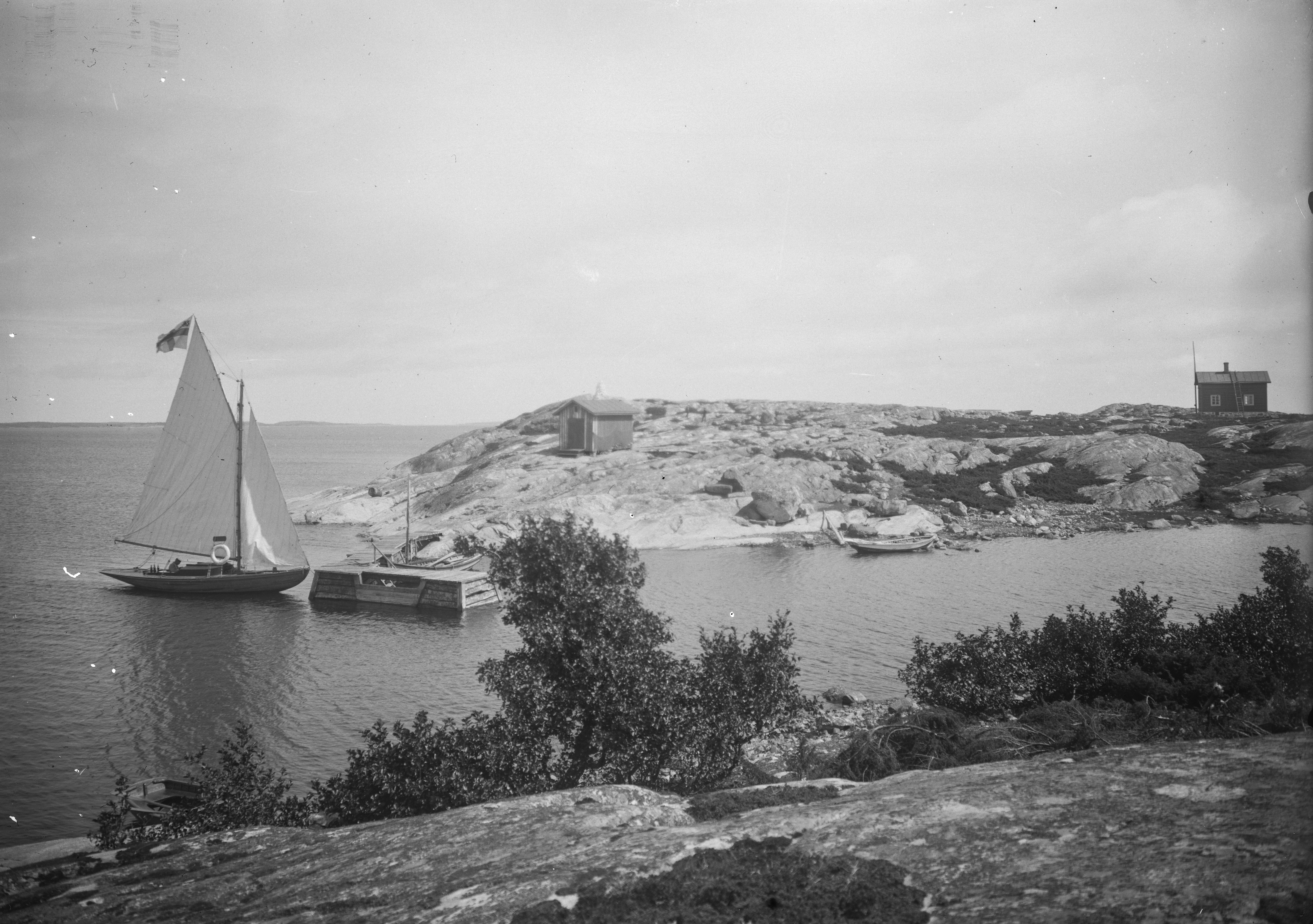 En gammal svartvit bil på en liten segelbåt som seglar in i en skärgårdsvik.