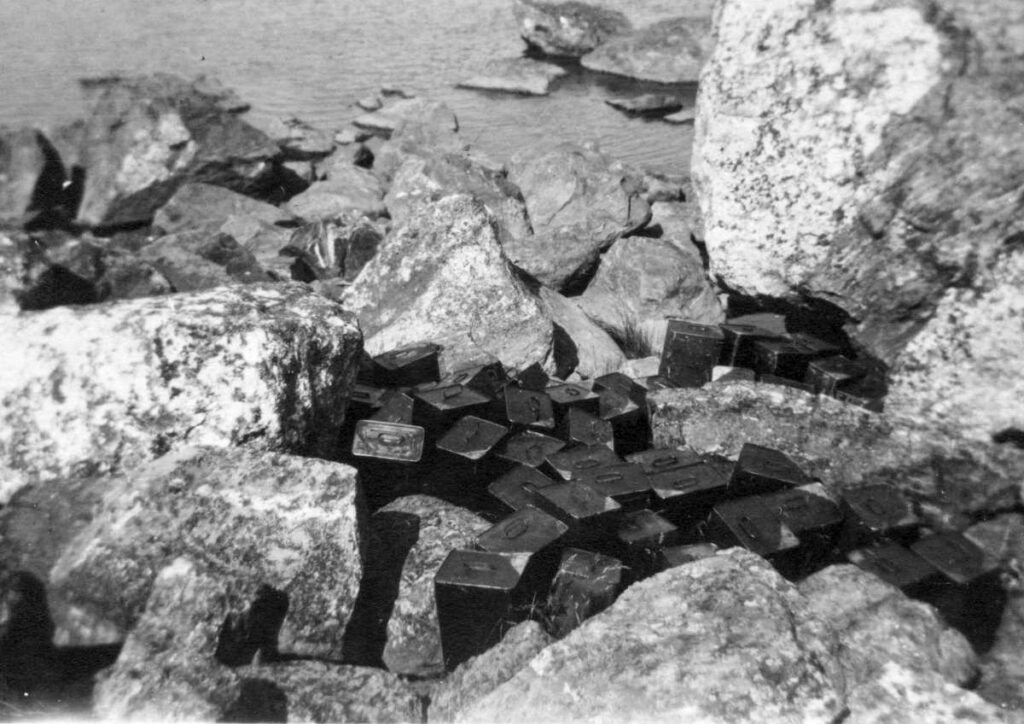 En gammal svartvit bild på stora stenar vid havsstranden, bland stenarna skymtar svarta kanistrar.