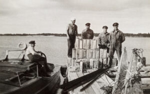 En gammal svartvit bild där fyra män sår på en brygga, framför sig har den en stor mängd gammaltida spritkanistrar.