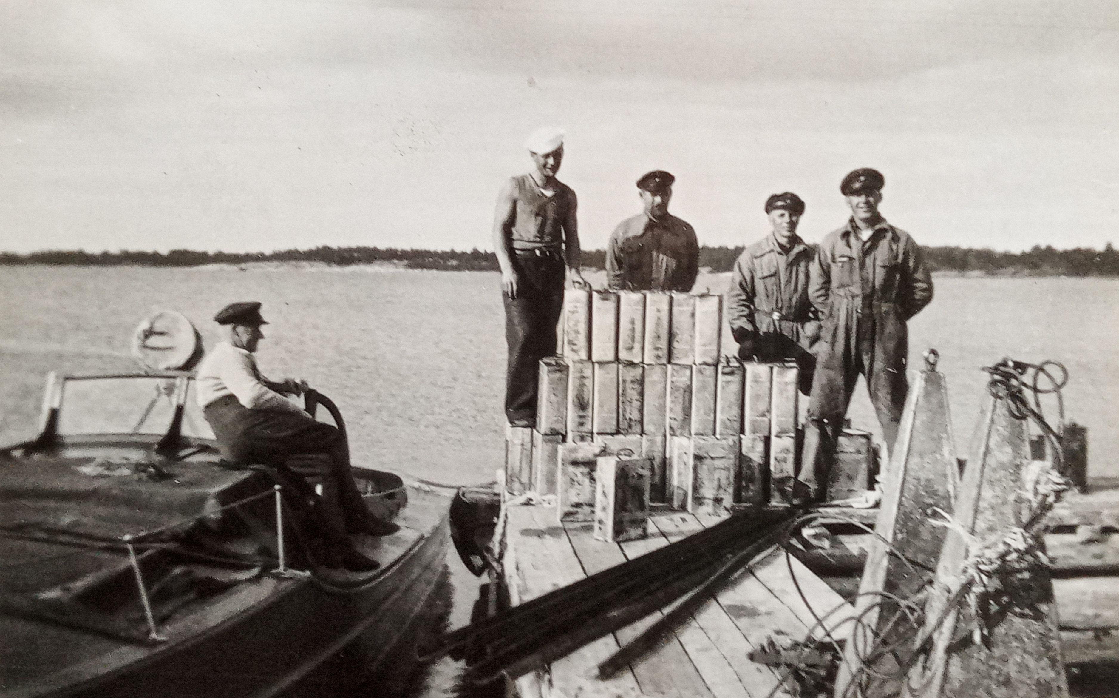En gammal svartvit bild där fyra män sår på en brygga, framför sig har den en stor mängd gammaltida spritkanistrar.