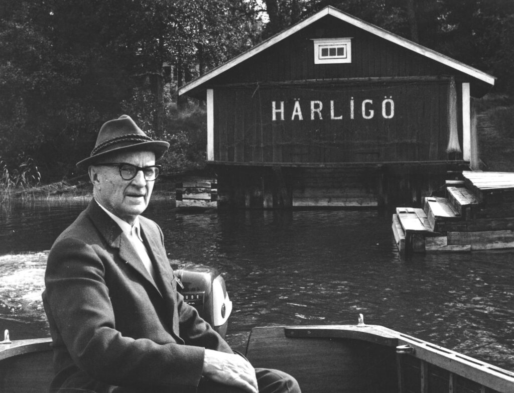 Svartvit bild på en man i kostym som kör aktersnurra. I bakgrunden ett båthus på vilket det står Härligö.