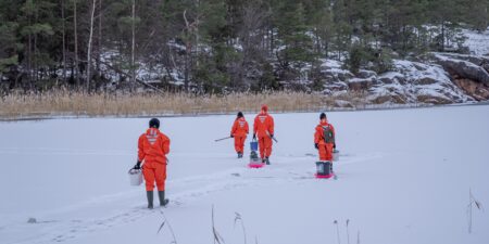 Människor i orangefärgade livräddningsdräkter vandrar på is med ämbar
