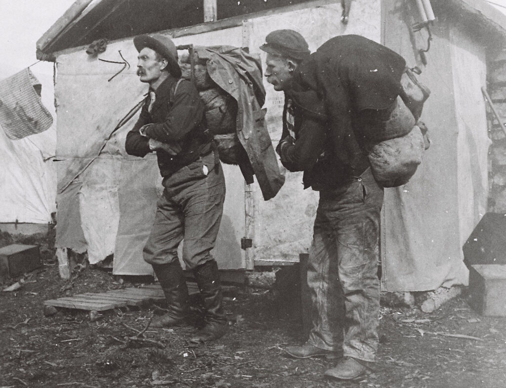 Gammal svart-vit bild på två män med ryggsäck på ryggen.