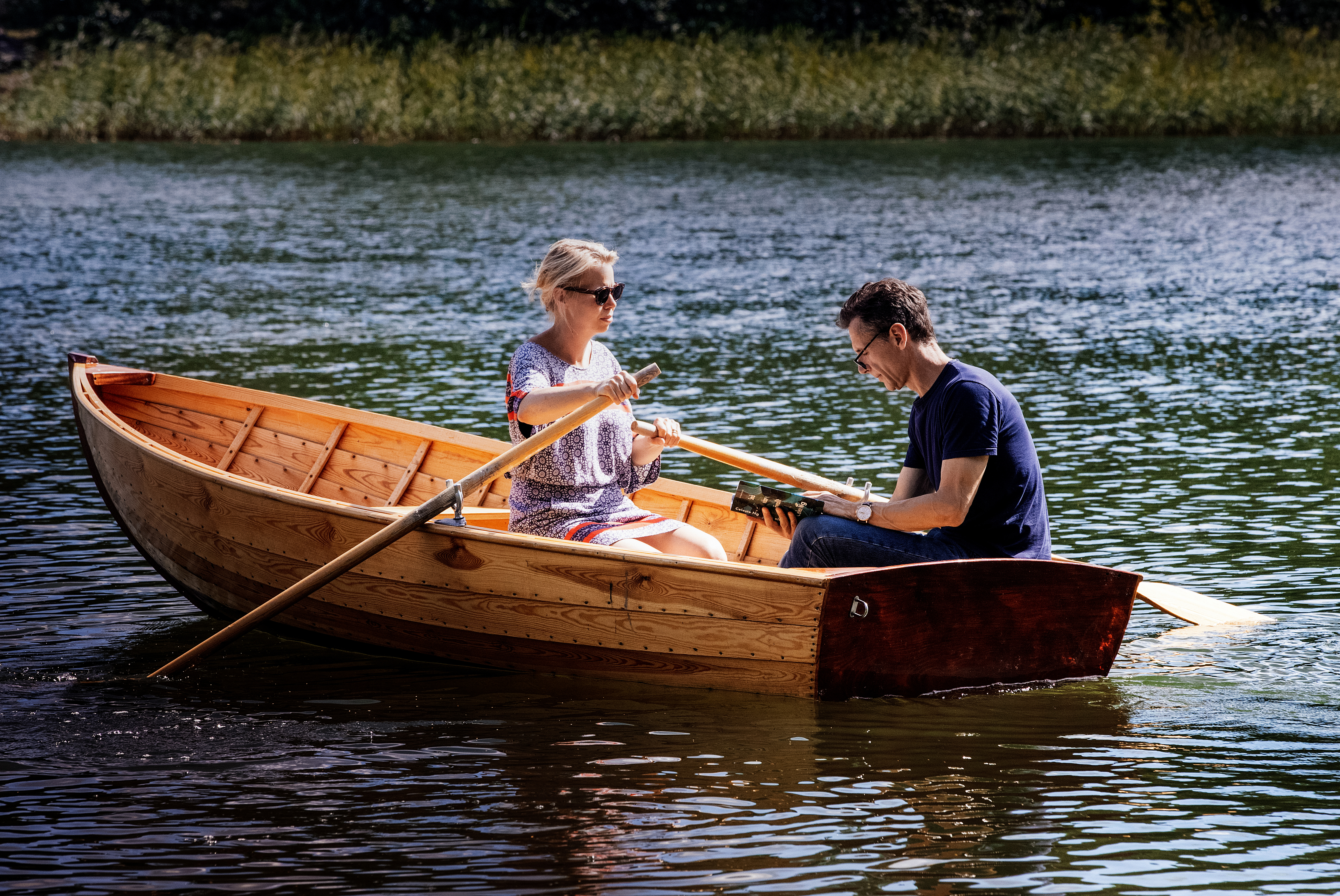 En kvinna och en man i en eka, kvinnan ror, mannen läser en bok.