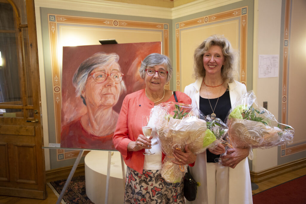 Kansler emerita Ulrika Wolf-Knuts och bildkonstnär Danelle Bergstrom med porträttet i bakgrunden och blombuketter i händerna. 