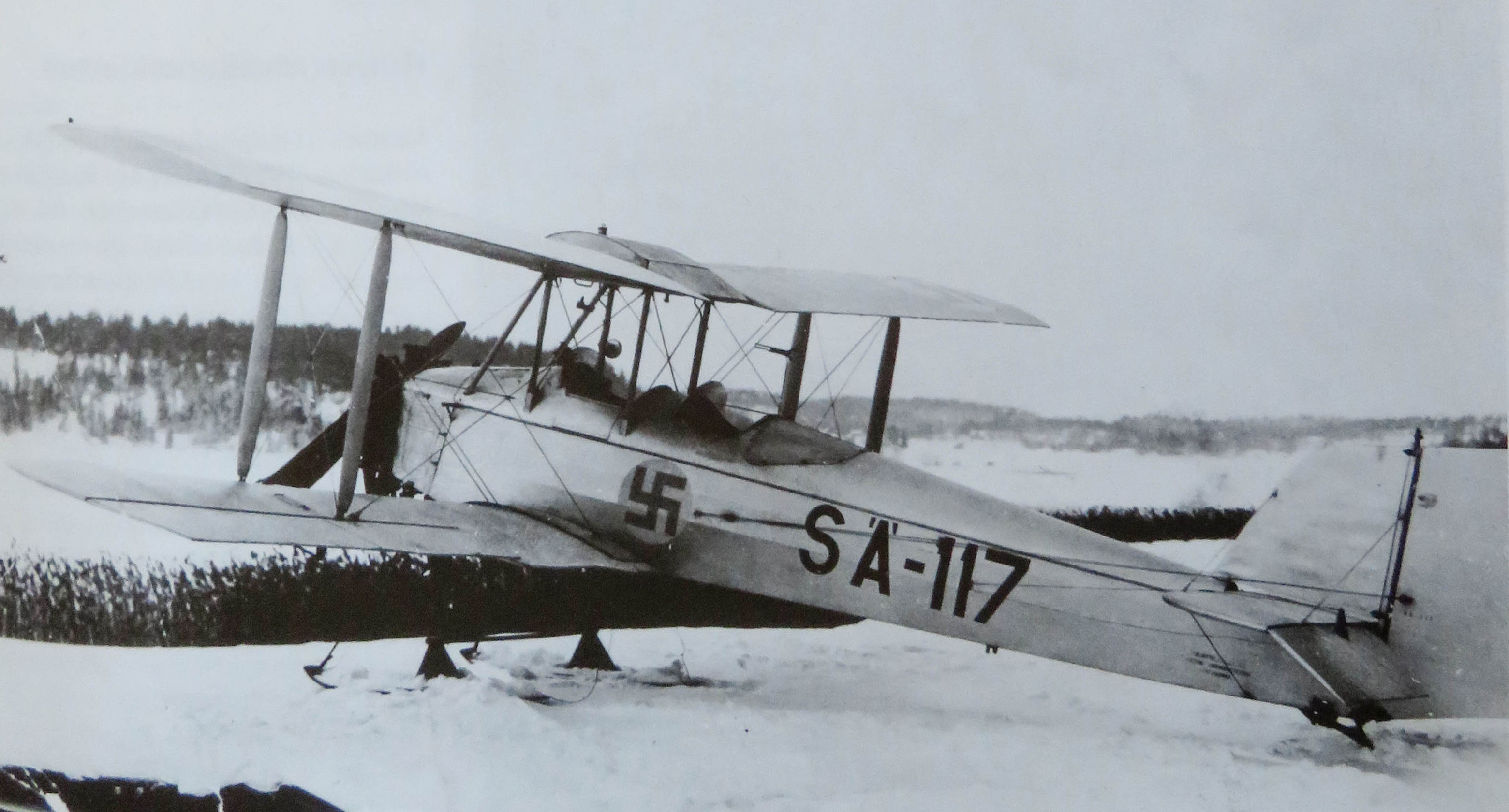 En svartvit bild på ett litet propellerplan som står på snöig mark.