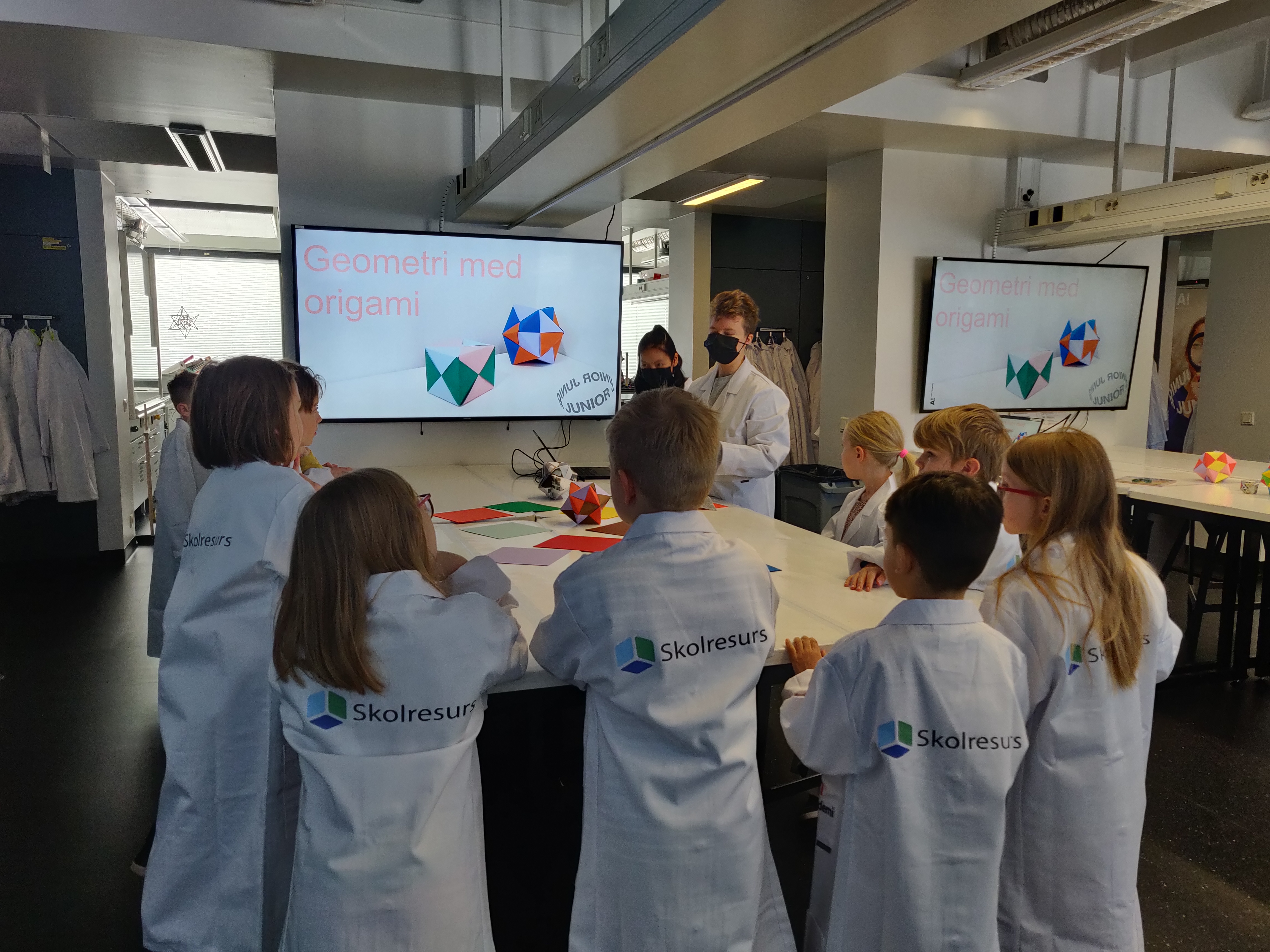 Barn i laborationsrockar med texten Skolresurs lyssnar på en presentation. På skärmen bakom presentatörerna står texten 