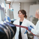 Två kvinnliga studerande i labbrockar gör mätningar vid en glovebox apparat i ett laboratorium.