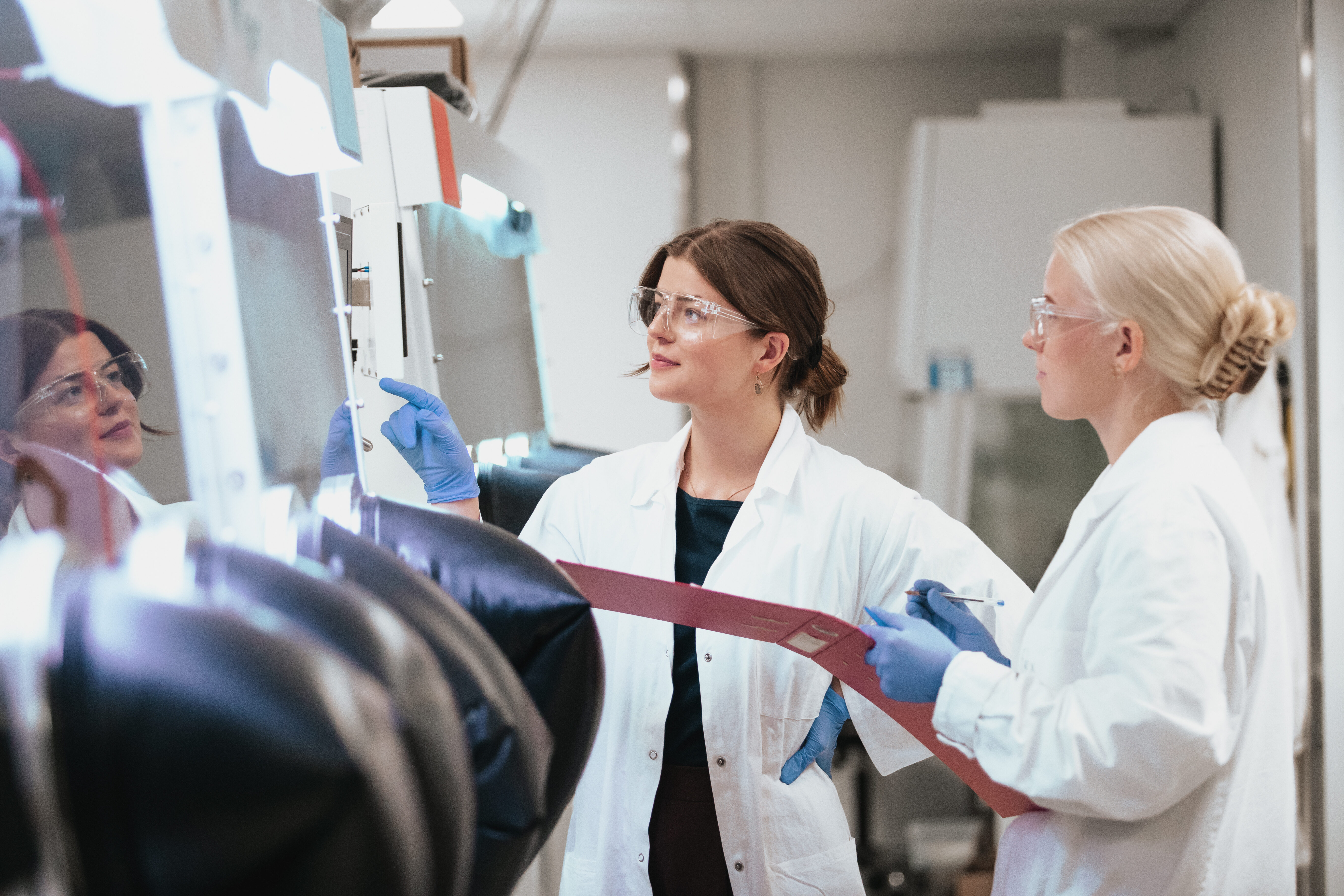 Två kvinnliga studerande i labbrockar gör mätningar vid en glovebox apparat i ett laboratorium.