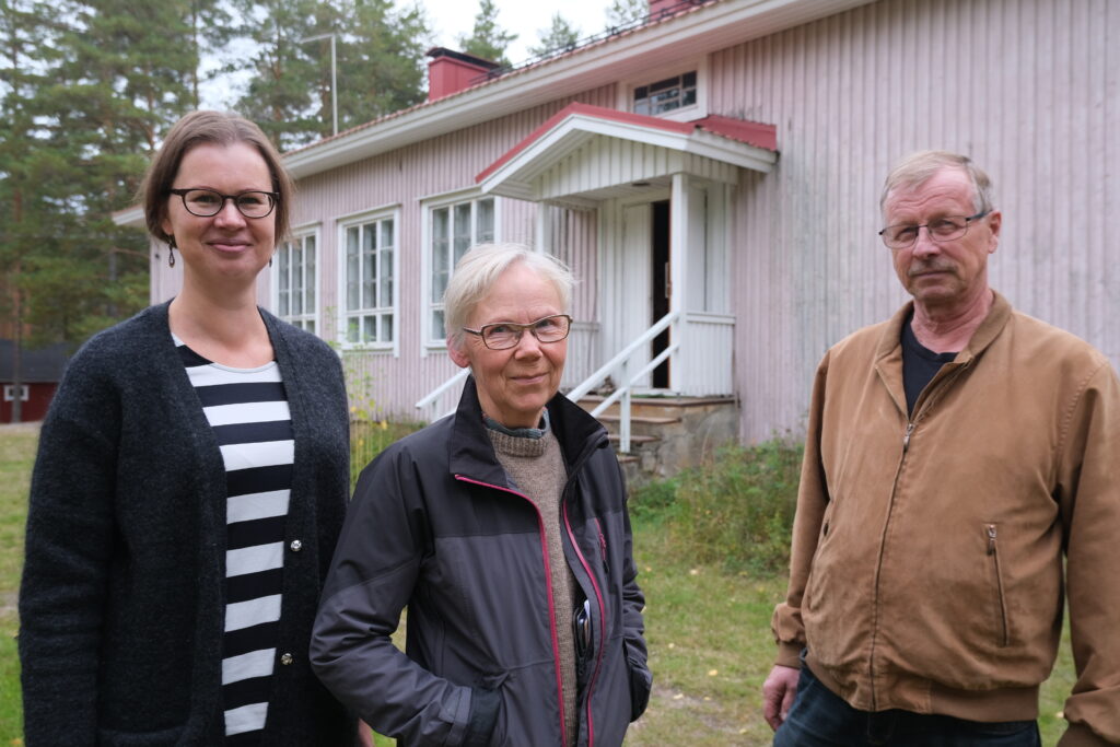 Tre leende personer står bredvid varandra, utomhus, framför en gammal byggnad.