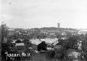 På bilden, från öst till väst, kan man se kyrkan till höger och i bakgrunden öns symbol, båken i granit, byggd 1862. Foto: Lauri Haapaniemi. Haapaniemi (1881–1946) arbetade på Aspö som lärare och präst under åren 1912–1919. Han förevigade öbornas liv med sin kamera och förekommer även själv på flera fotografier, vilket var möjligt med hjälp av en lång kamerautlösare. Lauri Haapaniemis bilder erbjuder en resa tillbaka i tiden till ölivet för över hundra år sedan.