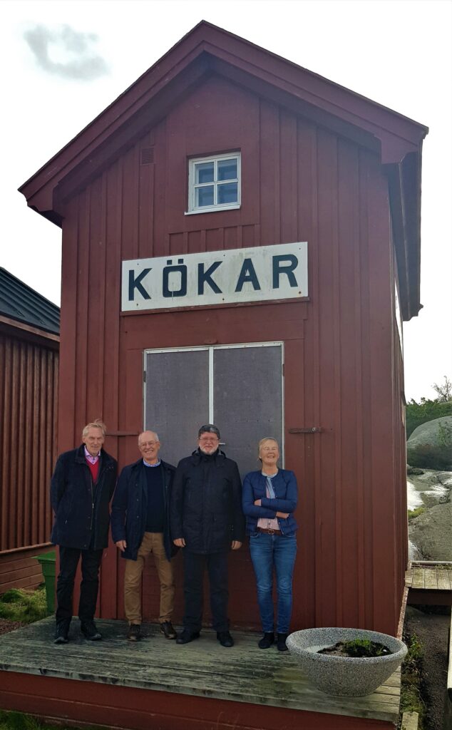 Fyra personer står framför ett rött hus. På husväggen en skylt med texten Kökar.