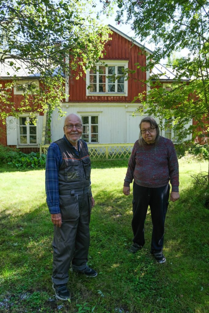 Två äldre män står på gräset framför ett röd-vitt hus.
