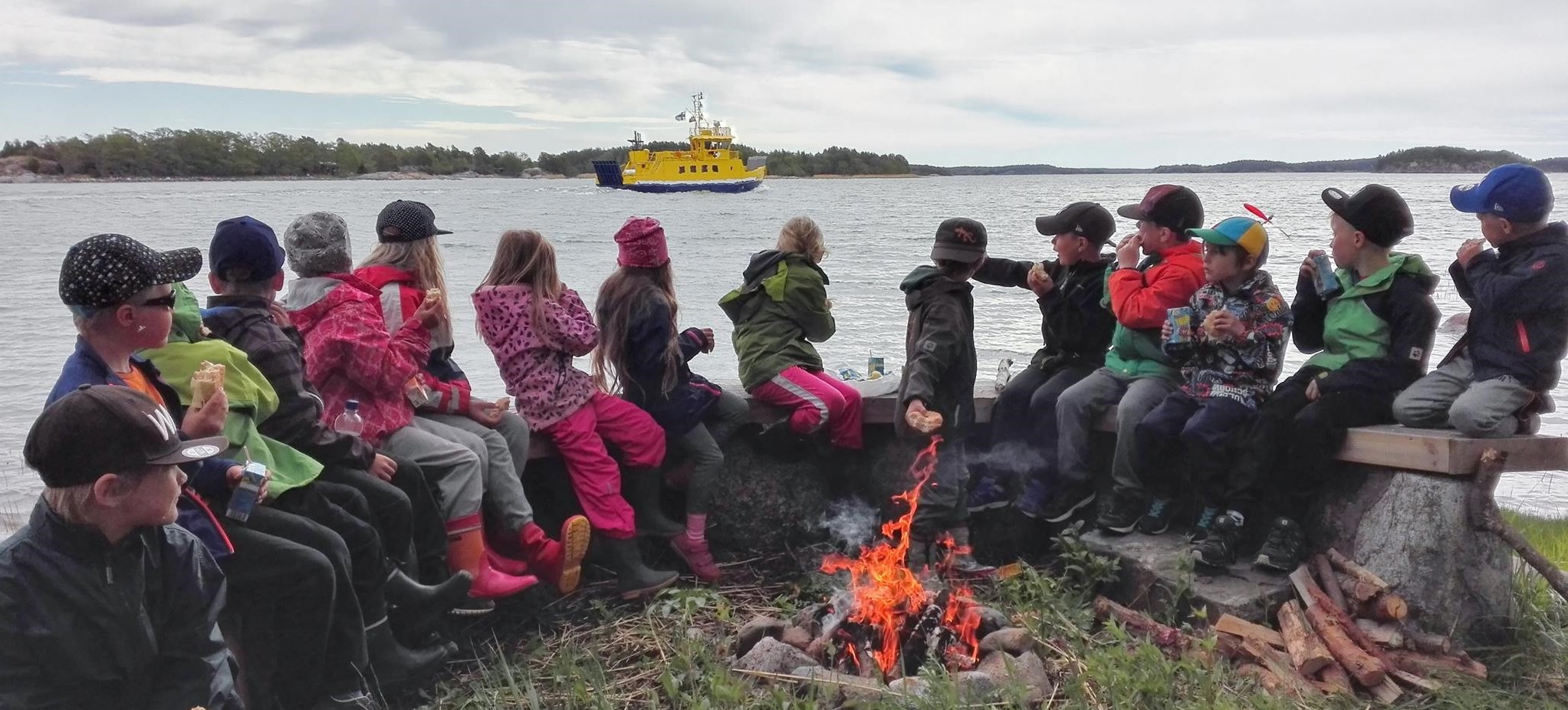 Barns sitter runt en lägereld, en färja åker förbi i bakgrunden.