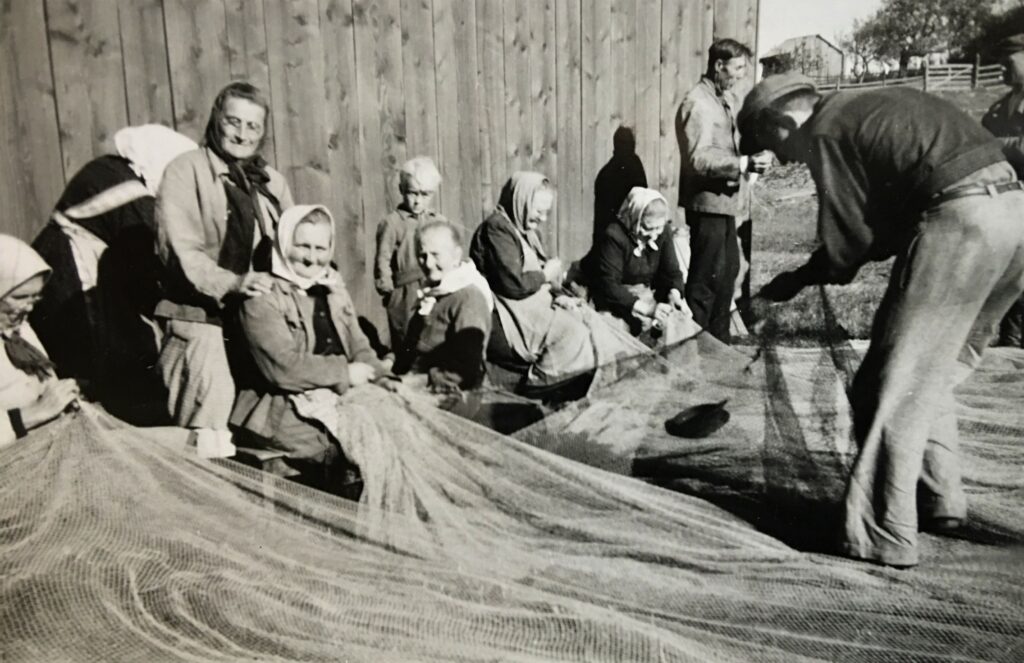 Svart-vit bild där en grupp människor sitter på rad med ett fisknät framför sig.