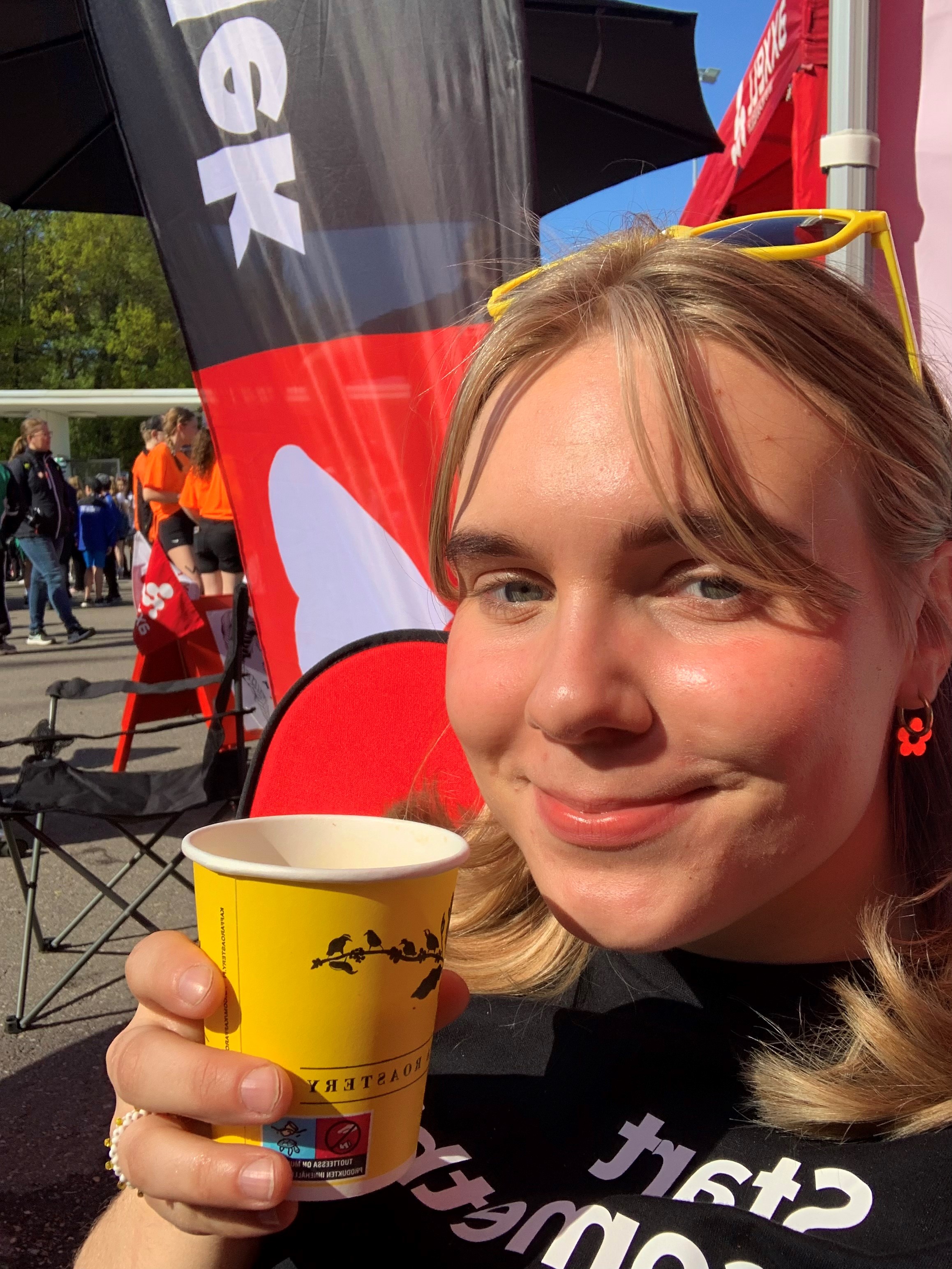 Porträtt av Elea som sitter i solen med en kaffe i handen och ler mot kameran