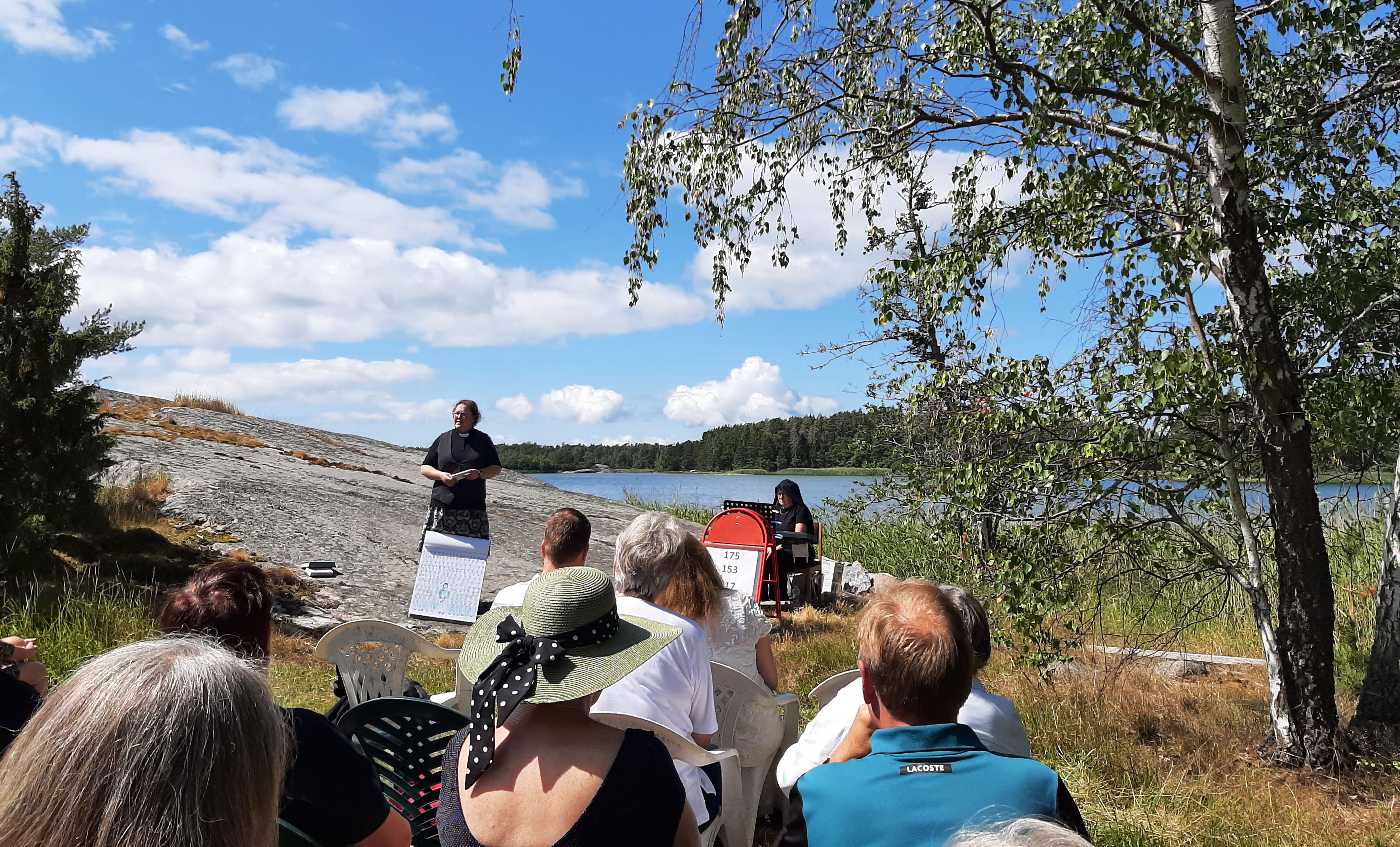 En utegudstjänst med församling i förgrunden och prästen och kantonr längre bort på en klippa.