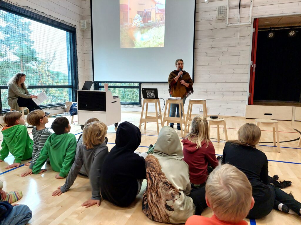 Rektor Päivi Soukka presenterar Velkua skolas verksamhet, en del av eleverna sitter längst fram på golvet och lyssnar.