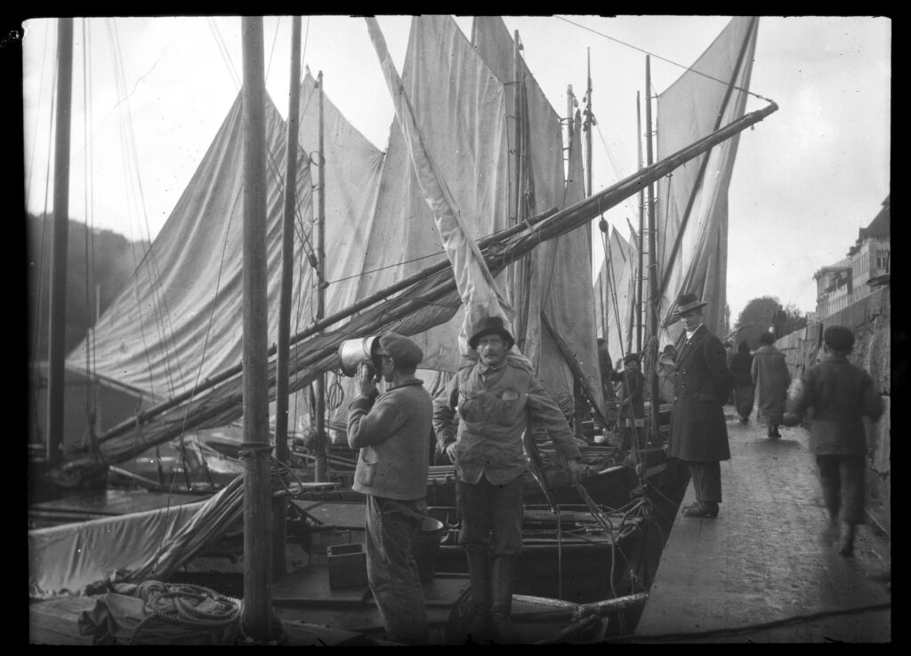 En svart-vit, gammal bild på segelbåtar förtöjda vid en kaj, några människor sysslar med sitt.