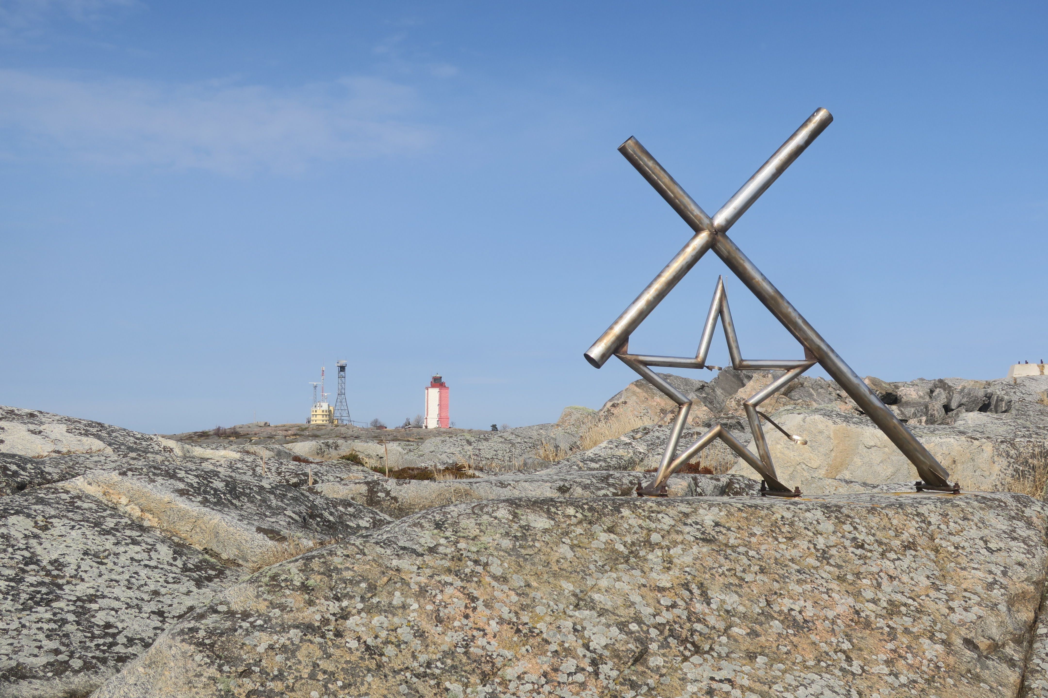 En stjärna och ett kors i metall står på ett berg. I bakgrunden syns fyren på Utö.