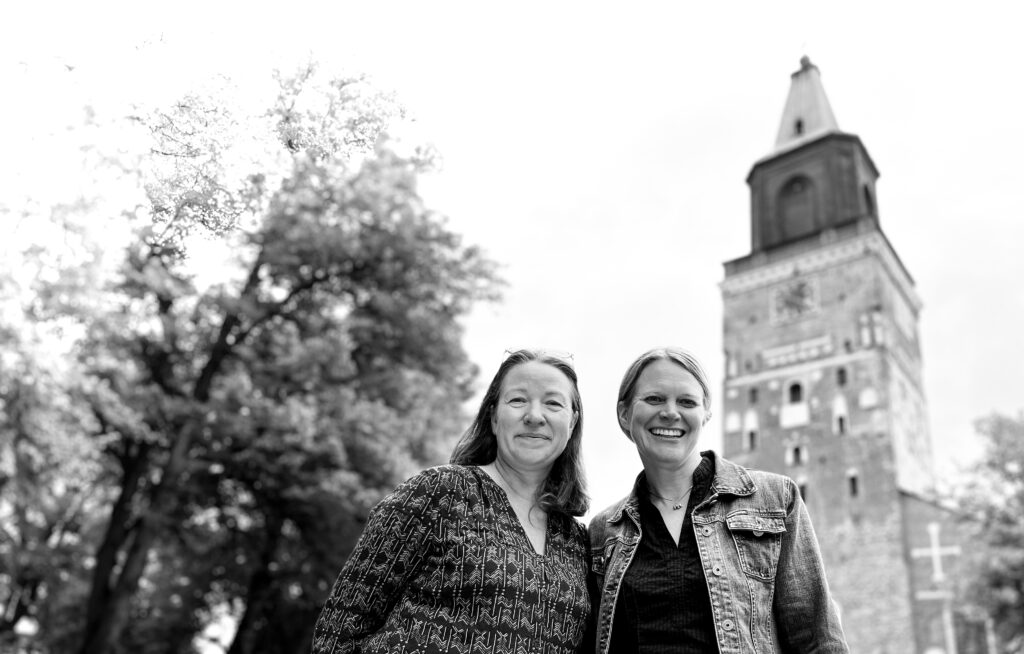 En svart-vit bild på två leende kvinnor framför Åbo somkyrkas höga torn.