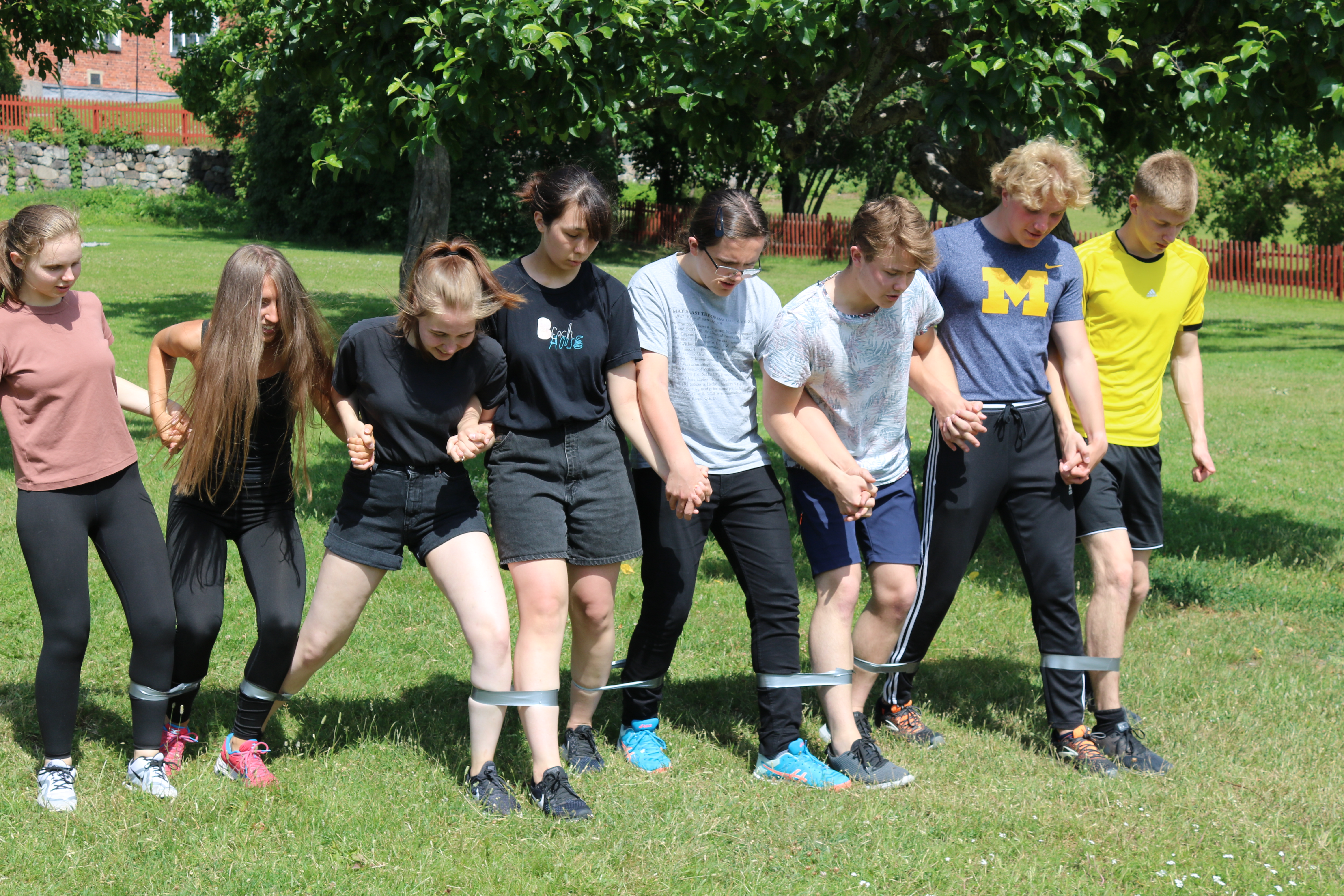 En grupp ungdomar som gör en samarbetsövning där de är fastbundna via vristerna till varandra och står på en gräsmatta.