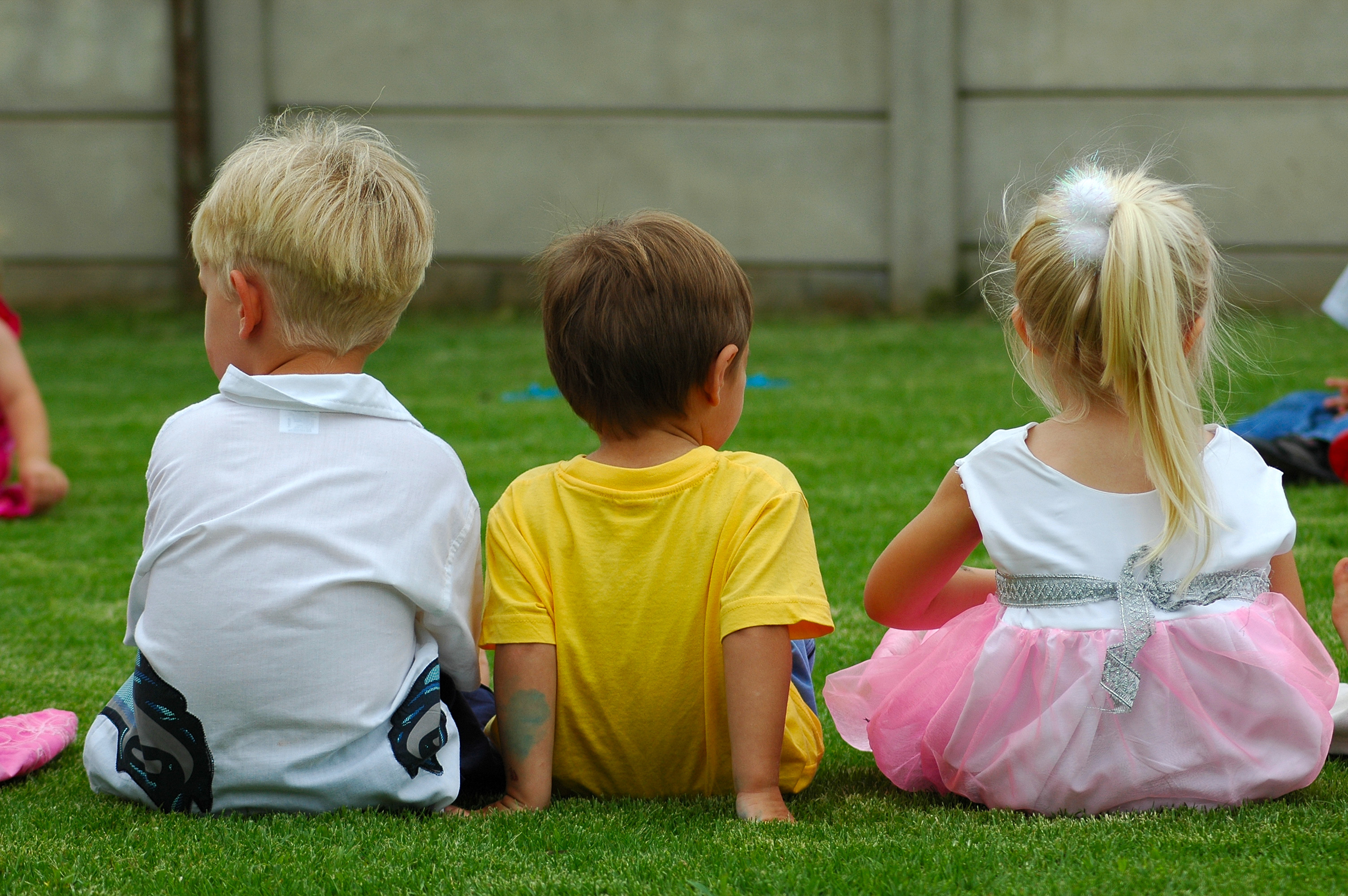 Tre små barn sitter på en gräsmatta med ryggarna mot kameran. Två pojkar och en flicka.