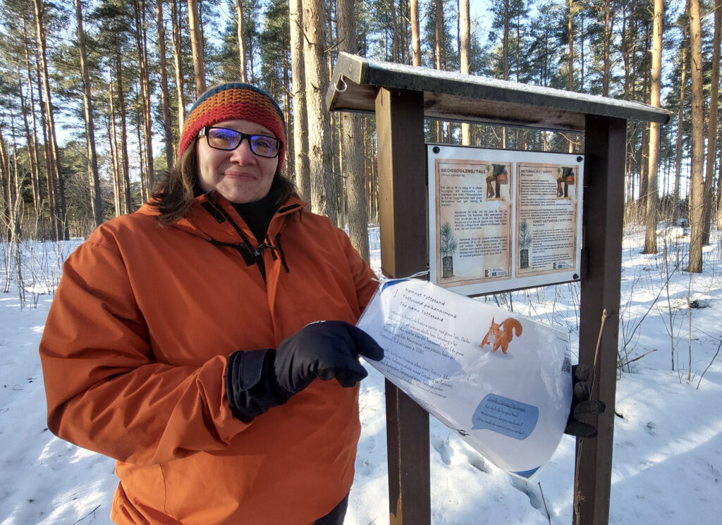 En leende kvinna ute i vintervädret vid en skylt med informationstext.
