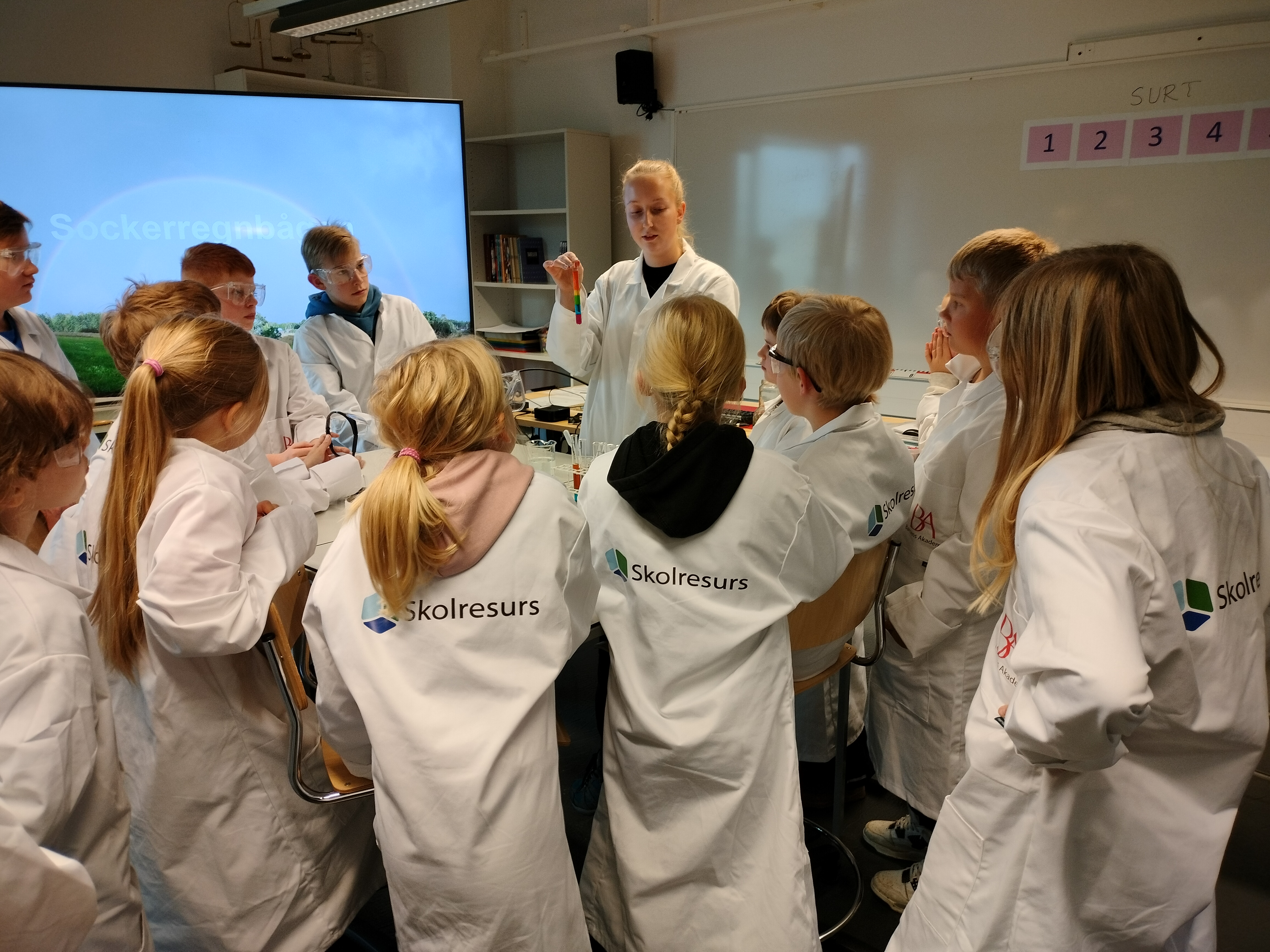 En grupp barn i vita labbrockar med Skolresurs logo är samlade runt ett bord och lyssnar på instruktioner. På skärmen i bakgrunden står det sockerregnbåge.