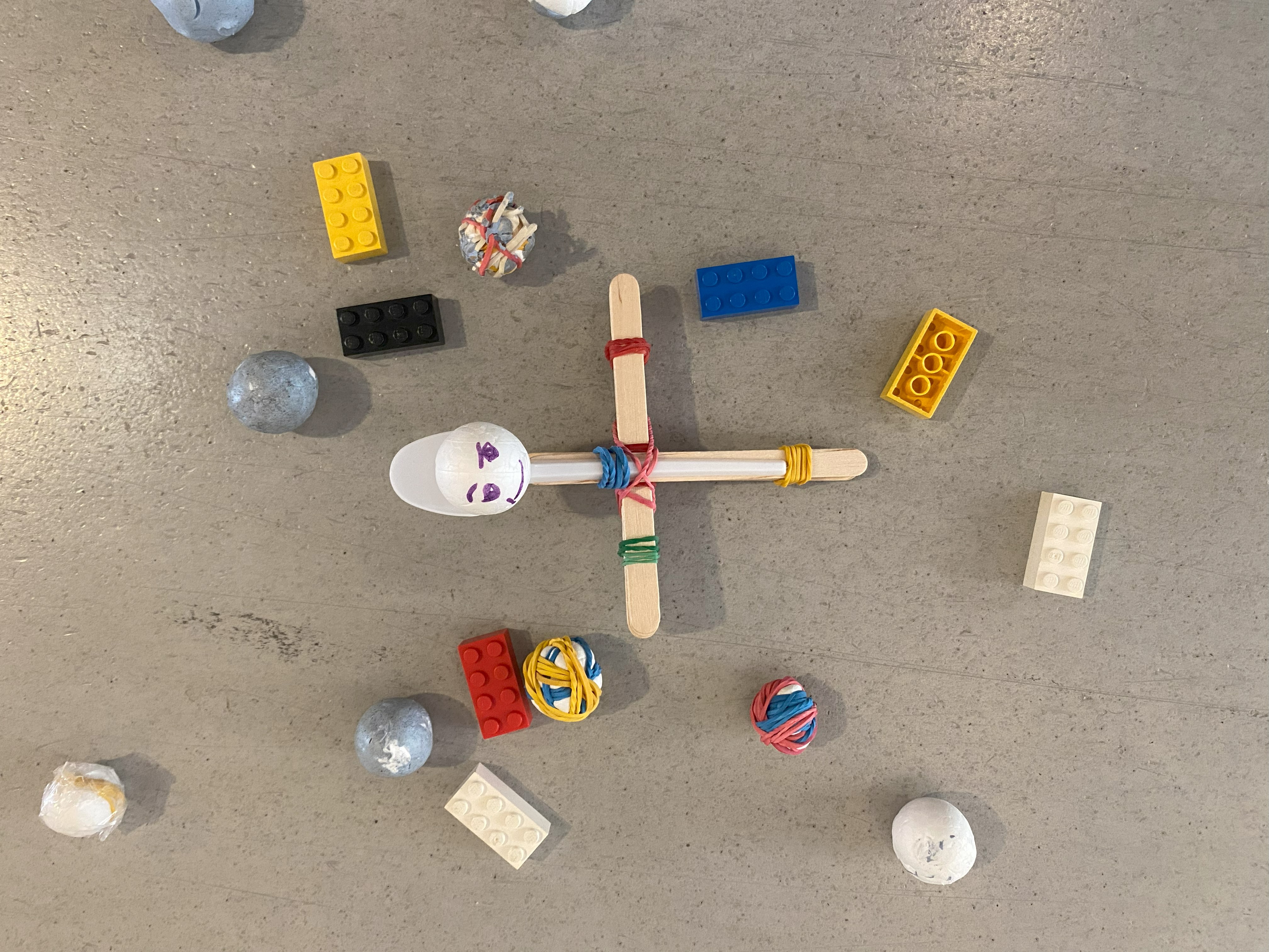 En katapult gjord av glasspinnar, gummiband och en sked med en styroxkula med pårittat ansikte, omgiven av kulor och legobitar.