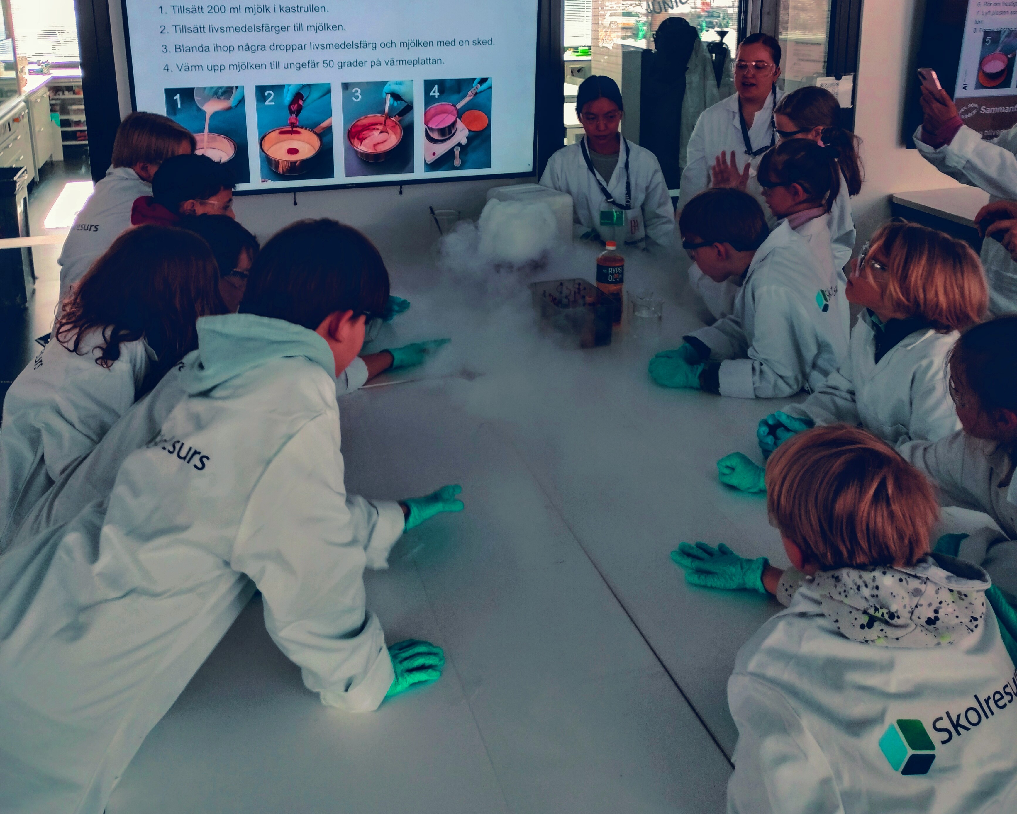 Bild från Barnens Akademis verkstad. Barn i labbrockar är samlade runt ett bord där de tittar på en rökformation. I bakgrunden syns recept för hur man framställer plast av mjölk.