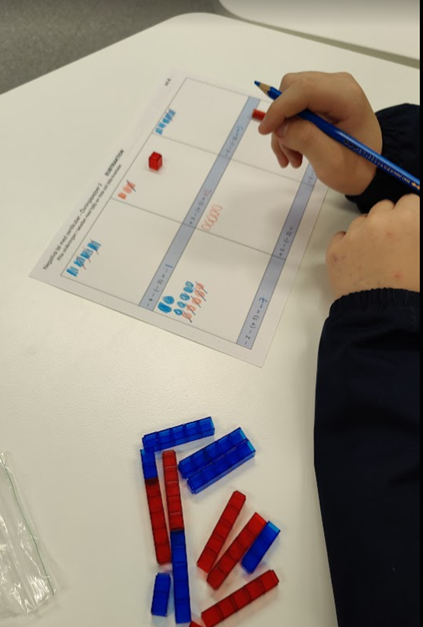 Närbild på elevs händer som håller i en penna och håller på och löser ett uppgiftspapper. På bordet finns blåa och röda klossar.
