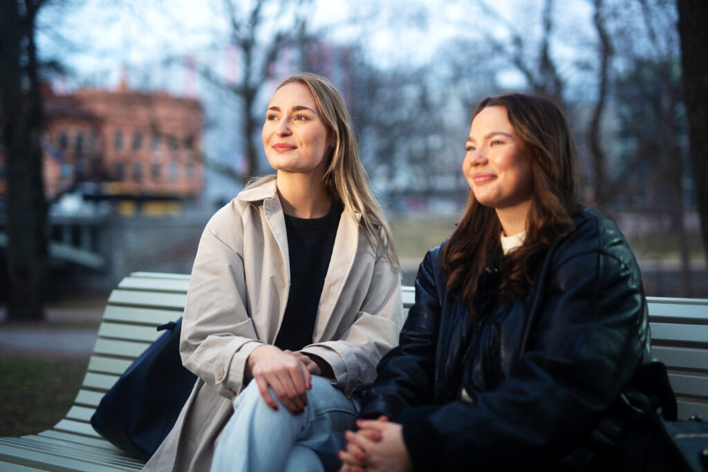 Två glada, unga kvinnor sitter på en bänk.