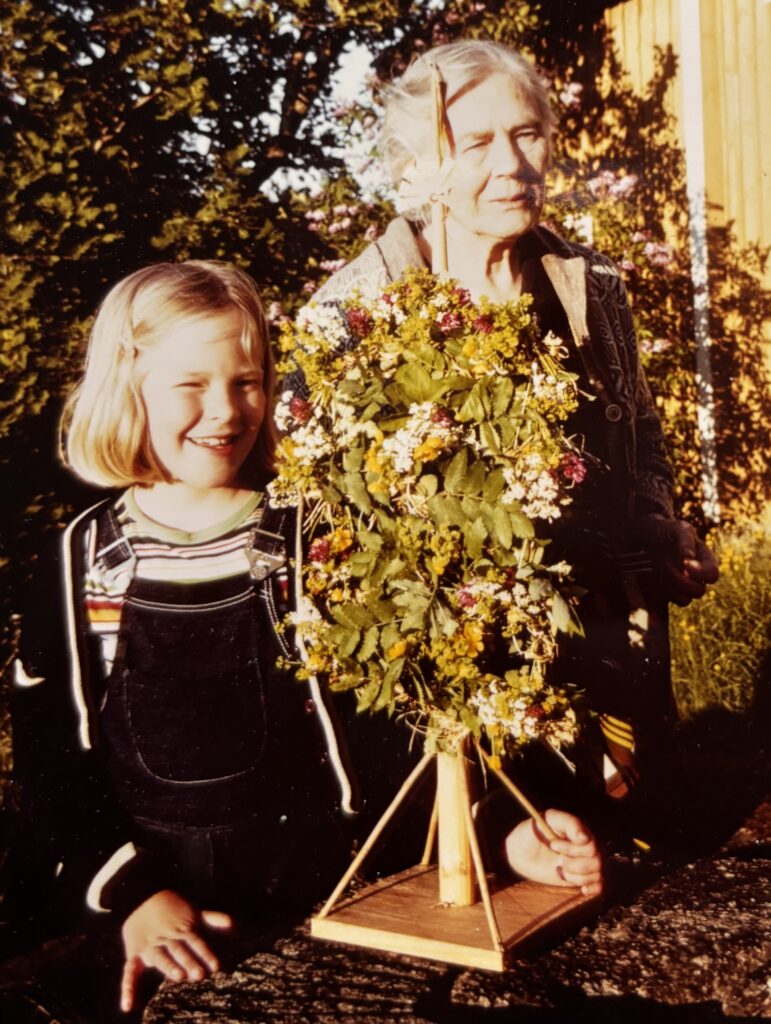 Cecilia Lundberg som barn, tillsammans med sin mormor. De står bakom en liten midsommarstång. 